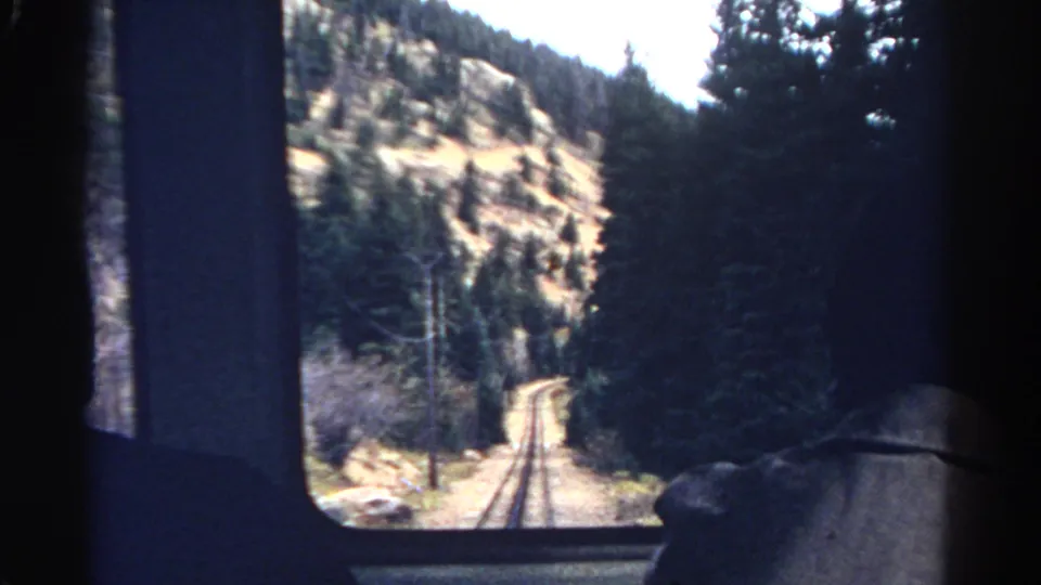 View from inside a moving 1967 Colorado train looking along tracks through pine trees, rocky slopes, and a bright mountain valley.