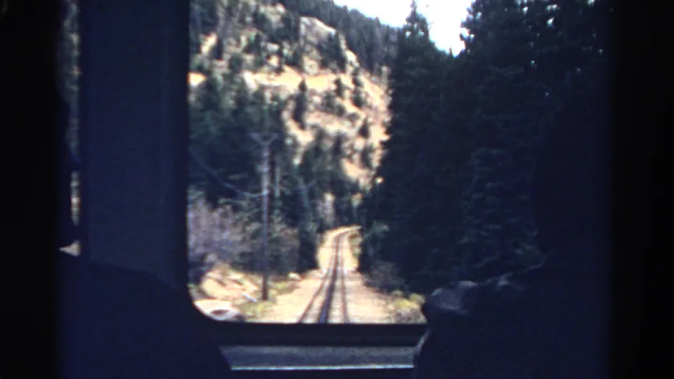A 1967 Colorado train window frames rails running through forest and rocky hills, with a passenger silhouette at the edge of the dim interior.