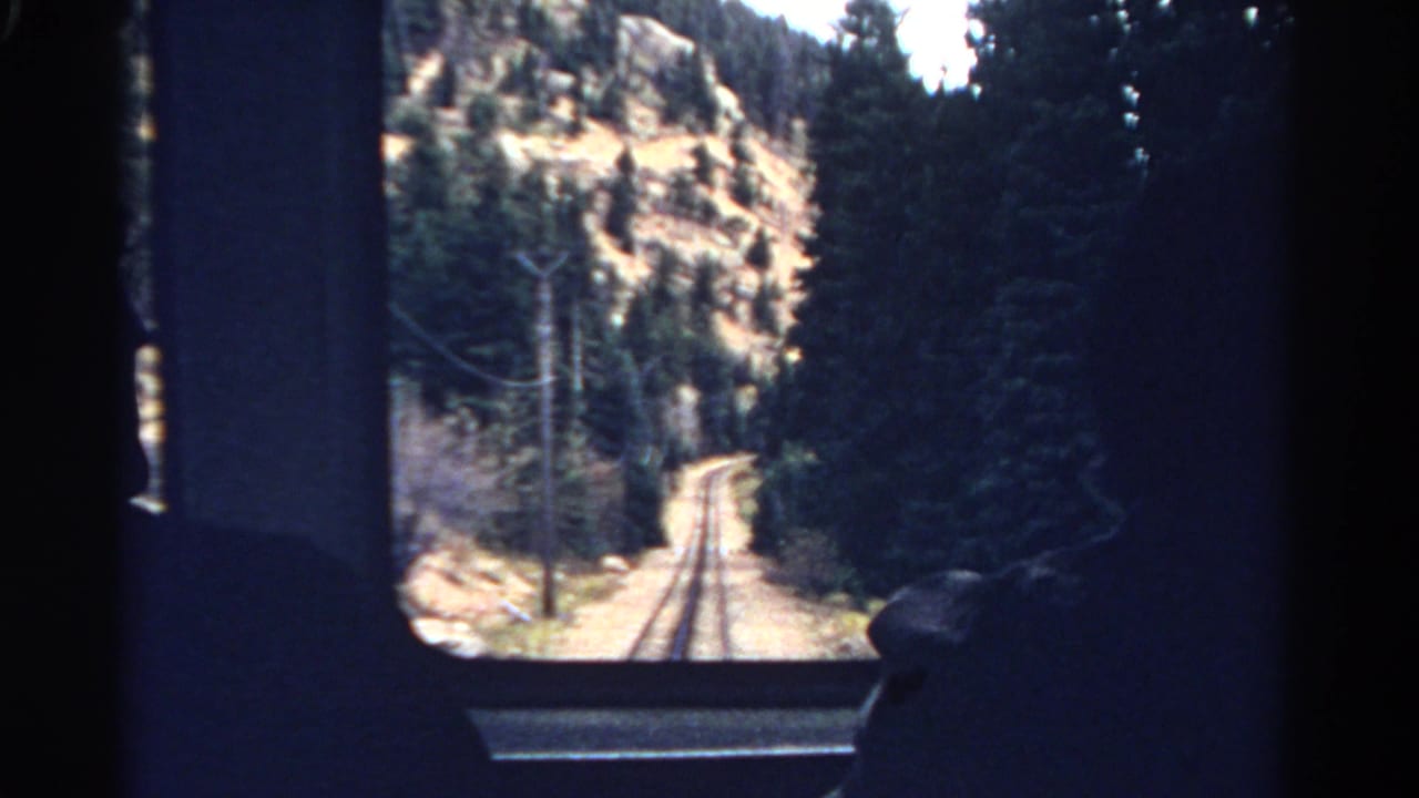 1967 Colorado train-window view of tracks entering pine forest below a dry rocky hillside, with passenger silhouettes at the edges.