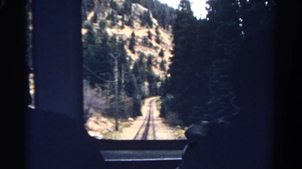 1967 Colorado train-window view of tracks entering pine forest below a dry rocky hillside, with passenger silhouettes at the edges.