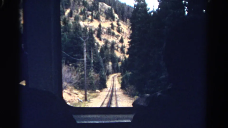 Colorado mountain rail view from inside a train, with track, pines, dry hillside, and a dark seated passenger silhouette.