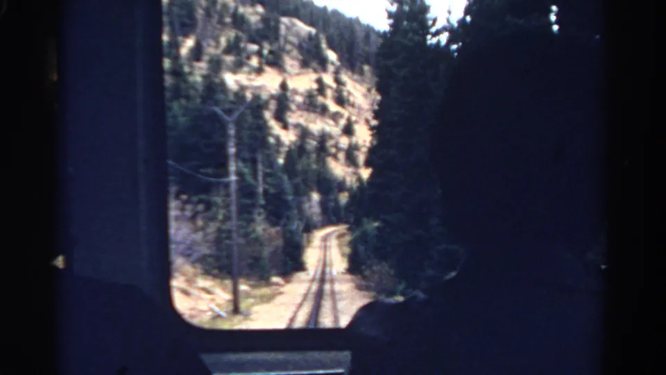 Home-movie view from a 1967 Colorado train ride, looking past a window frame toward rails, pines, and a dry mountain slope.