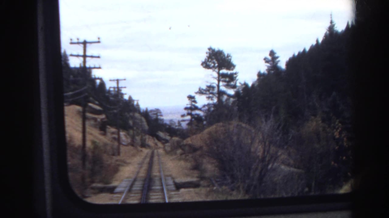 Passenger-window view of Colorado mountain rail tracks in 1967, framed by utility poles, pine trees, brush, and a cloudy sky.