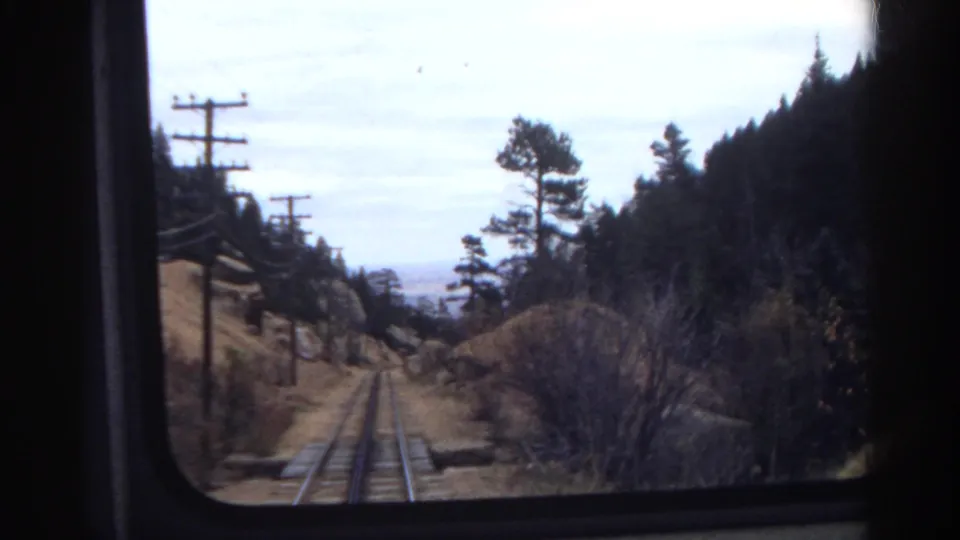 Passenger-window view of Colorado mountain rail tracks in 1967, framed by utility poles, pine trees, brush, and a cloudy sky.