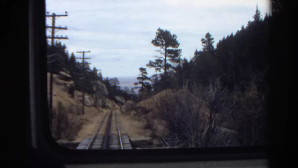 1967 Colorado 8mm train-window view showing rail tracks, roadside utility poles, pine trees, brush, and distant haze.