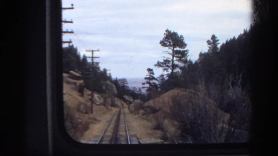 Colorado passenger-train view from 1967, with tracks leading through pine trees, brush, utility poles, and dark window edges.