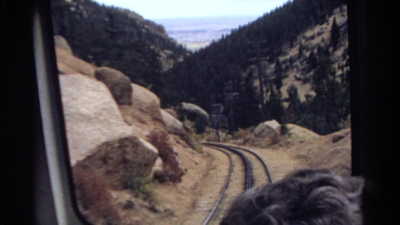 Passenger-window view of a Colorado mountain rail line curving past boulders and dark pine slopes in 1967 footage.