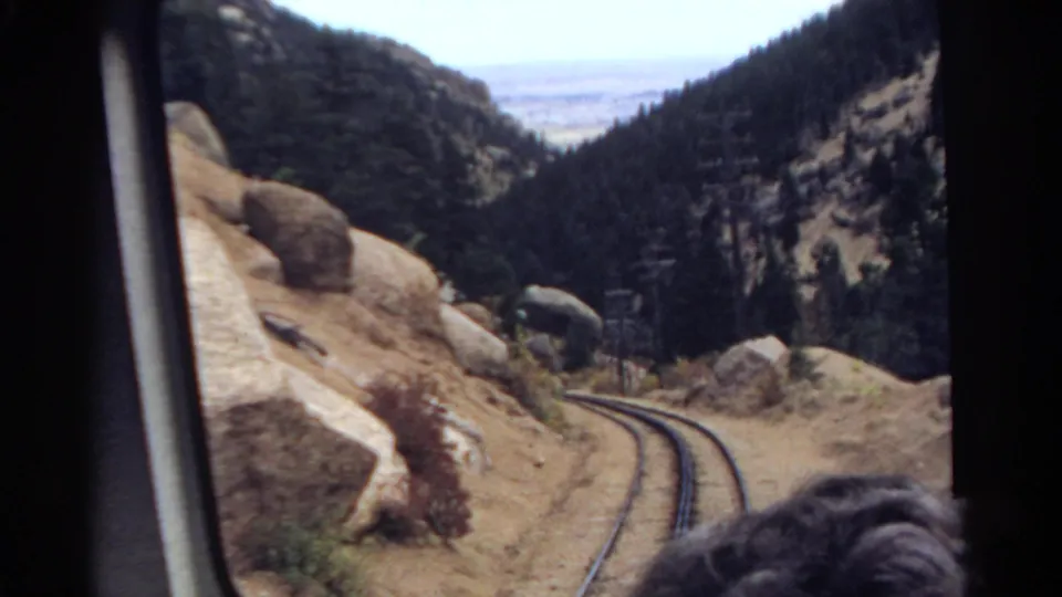 1967 Colorado home-movie frame showing rails dropping through a mountain pass with boulders, pines, and a hazy plain beyond.