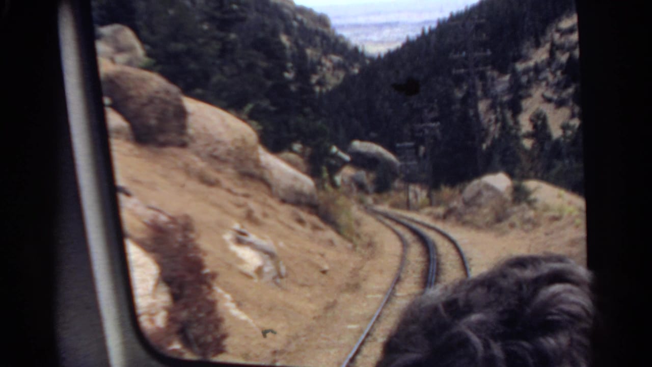 1967 Colorado home-movie frame looking from inside a train toward curving rails, rocky slopes, pine forest, and a passenger's hair in the foreground.