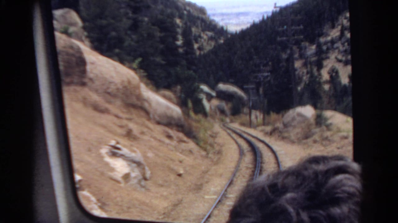 From inside a 1967 Colorado train, the frame shows rails bending through a rocky mountain pass with pine trees and a passenger partly visible at the window.