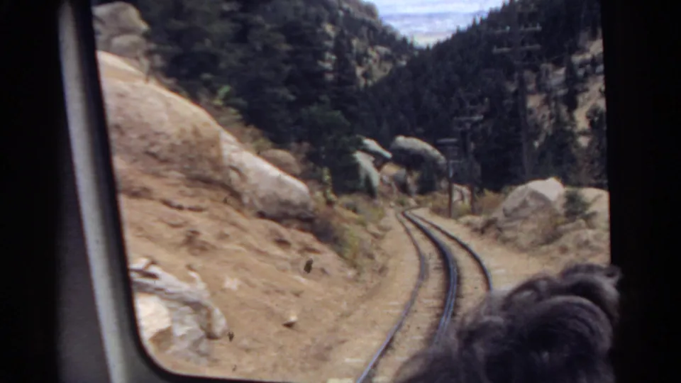 1967 Colorado 8mm frame showing a passenger-window view of railway tracks running between rocky slopes and dark pine forest.