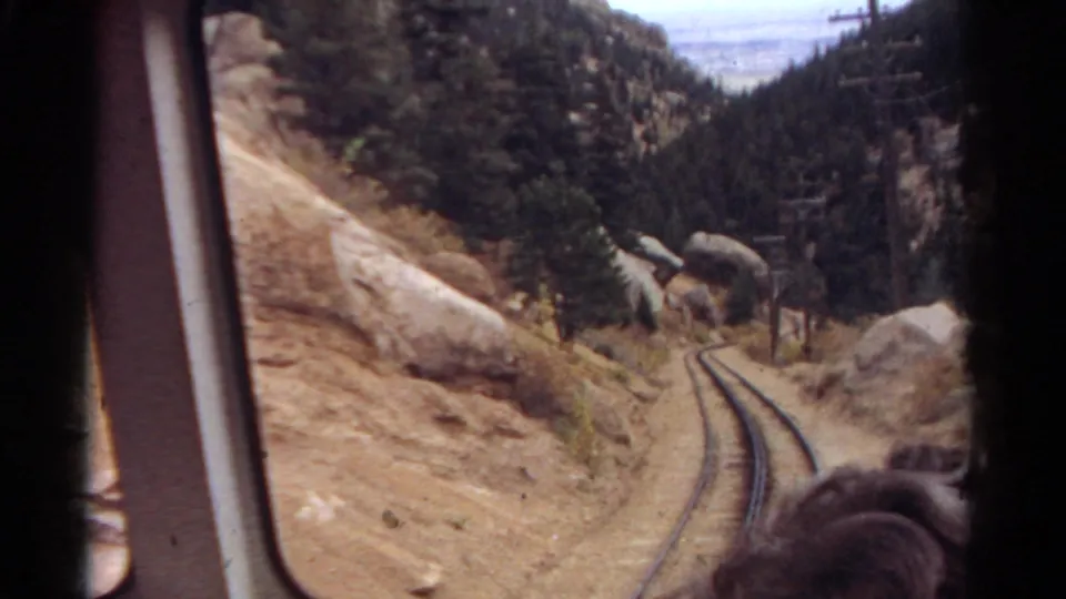 The final 1967 Colorado frame looks out from a train window at curving rails, dry rocky ground, boulders, pine forest, and a partial passenger foreground.