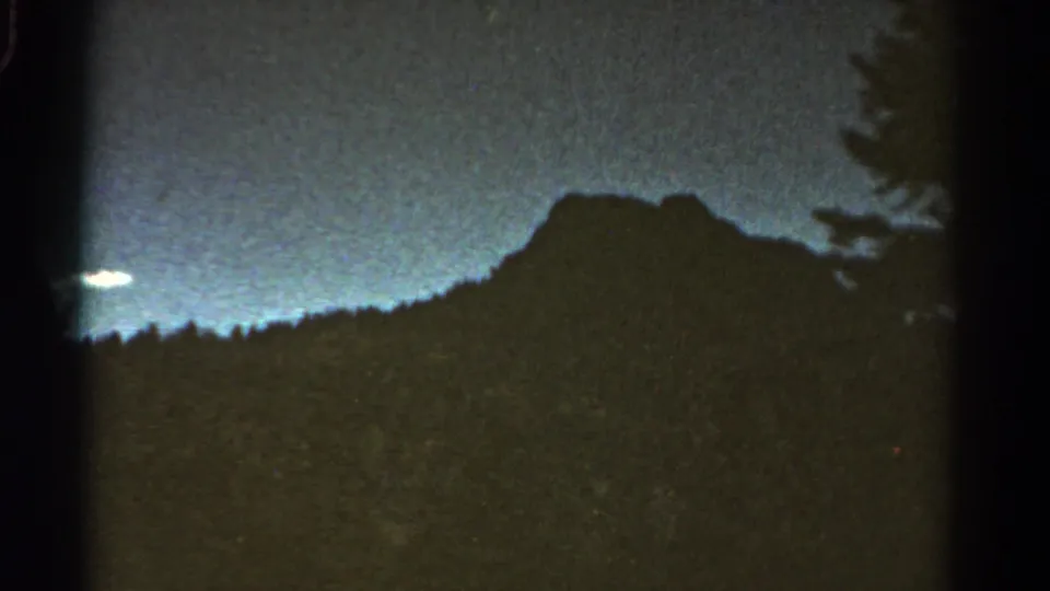 1967 Colorado 8mm frame showing a dark mountain form and evergreen tree edge beneath a dim blue sky.
