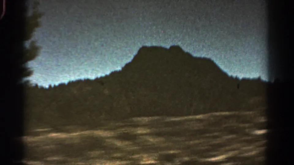 1967 Colorado road-trip frame with a dark mountain peak, treeline, pale dusk sky, and blurred roadside foreground.