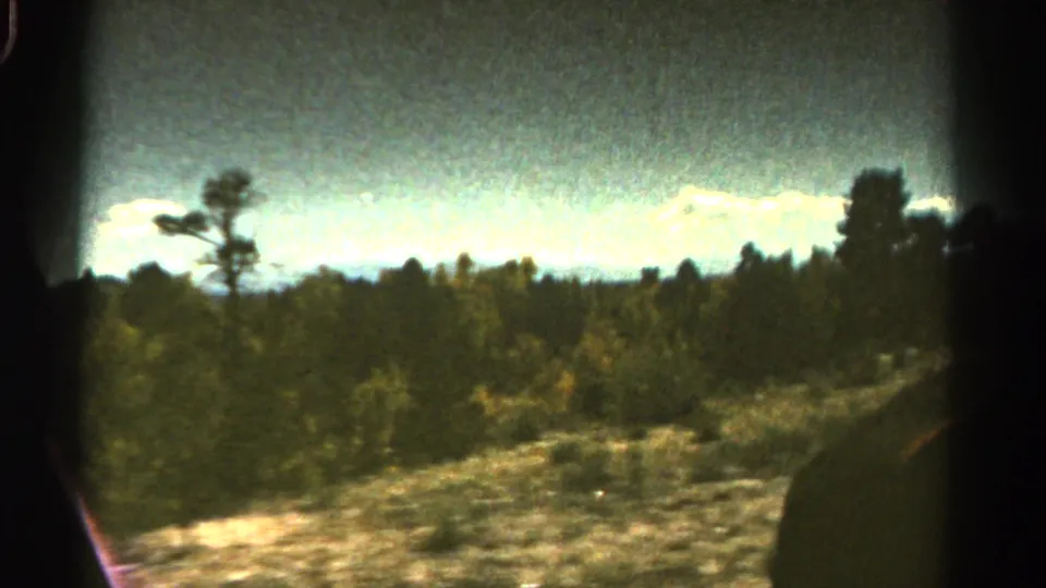 1967 Colorado roadside view with trees, distant mountains, and blue sky, framed by dark vehicle or film edges.