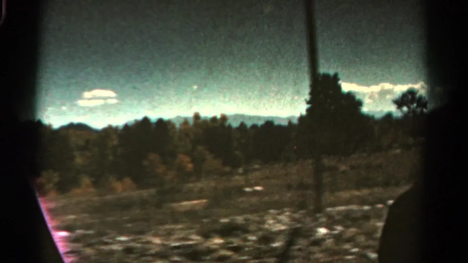 Wide 8mm Colorado landscape frame with tree line, distant mountains, roadside brush, clouds, and a dark vertical edge.