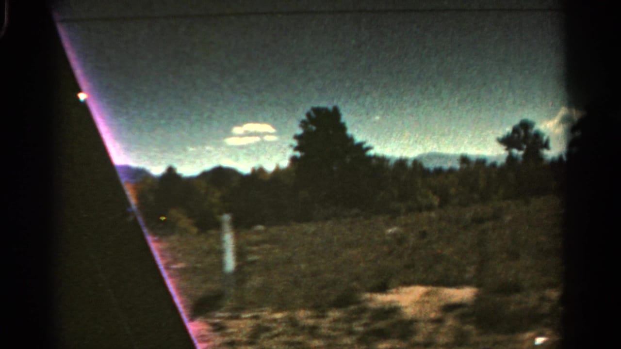 1967 Colorado road-travel frame with a pale roadside post, scrub terrain, trees, distant peaks, clouds, and heavy 8mm frame edging.