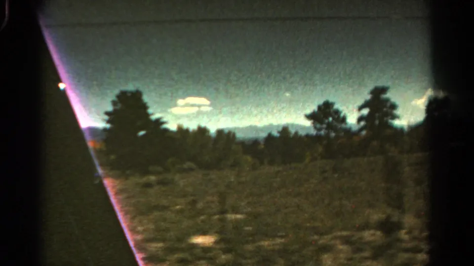 1967 Colorado 8mm frame showing low vegetation, dark trees, distant mountains, bright clouds, and prominent magenta lens flare at the edge.