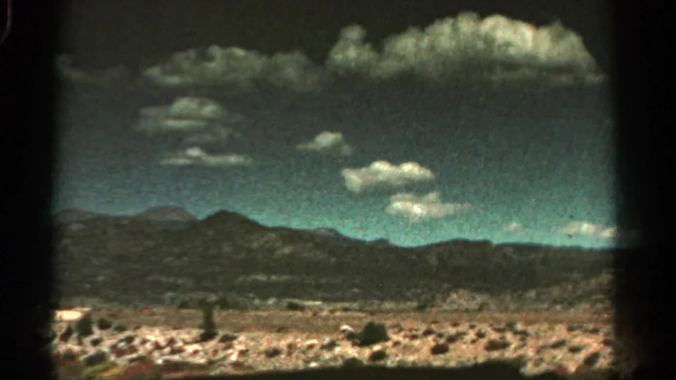 1967 Colorado 8mm frame of a broader rocky landscape, low mountain ridge, textured sky, and clustered white clouds.