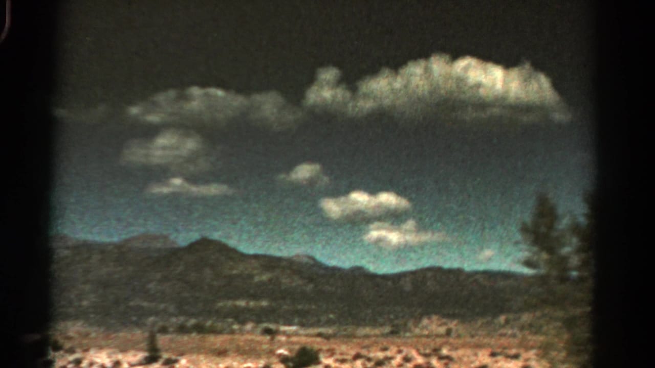 1967 Colorado 8mm frame showing distant desert mountains, scrubby terrain, and scattered clouds seen from a moving roadside view.