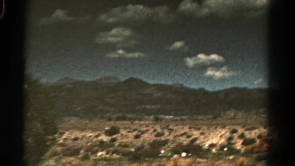 1967 Colorado 8mm frame of open rocky terrain and low mountain ridges under scattered bright clouds.