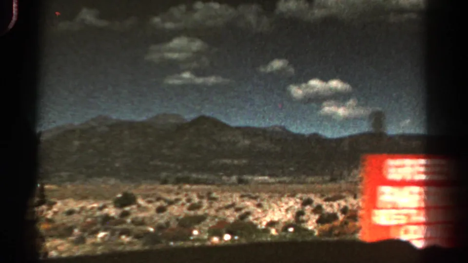 1967 Colorado roadside frame with desert mountains, scattered clouds, and a partially visible red Green Parrot Restaurant sign at right.