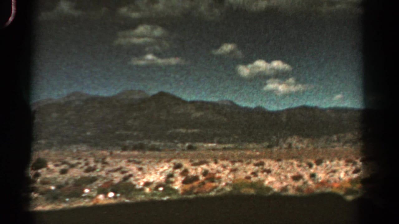 1967 Colorado home-movie frame from a moving vehicle, with dry open ground, distant mountains, and small clouds in a blue sky.