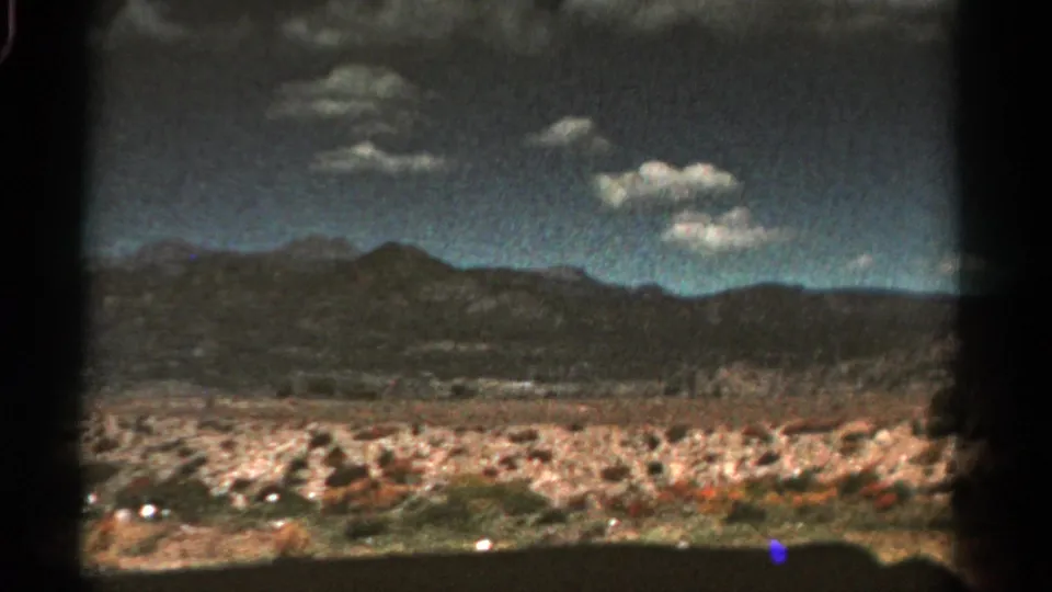 1967 Colorado 8mm frame showing dry roadside vegetation, distant mountains, scattered clouds, and visible film-gate edges.