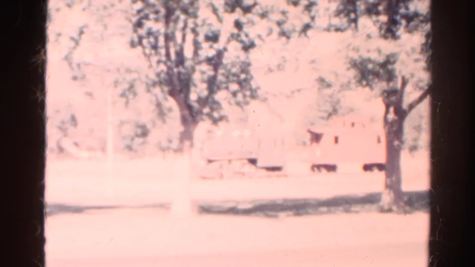 1967 Colorado 8mm frame with pink color shift, mature trees, pale open ground, and a small background rail-car-like object.