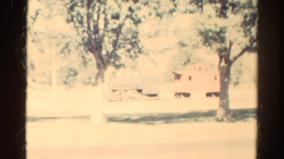 1967 Colorado 8mm frame showing a washed-out rural rail setting with trees, track, and a faint red car or caboose beyond the trunks.