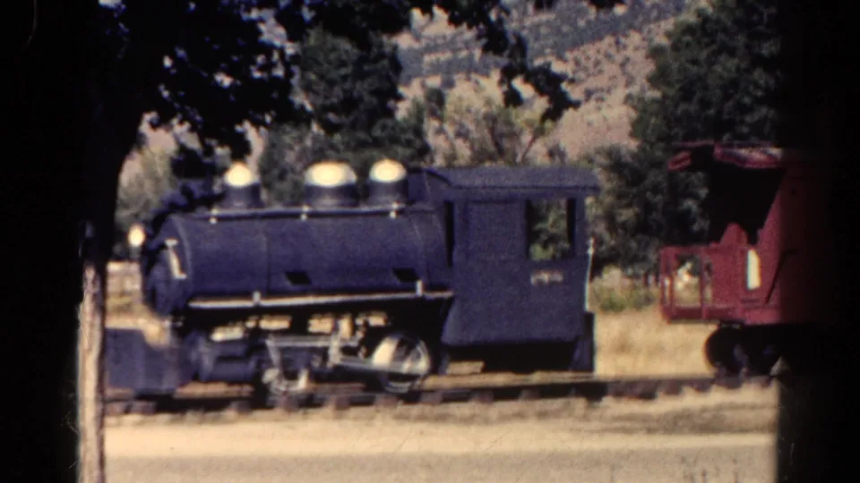 1967 Colorado home-movie frame with a dark blue steam-style locomotive on rails and a red caboose partly visible to the right.