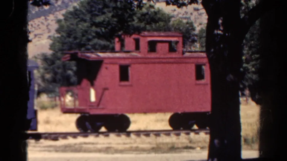1967 Colorado home-movie frame focused on a red caboose parked on track, with trees, dry grass, and hills behind it.