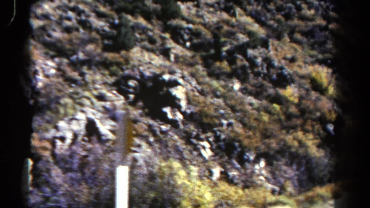 1967 Colorado travel frame with rugged hillside, scrub growth and vertical roadside posts viewed from inside a bus.