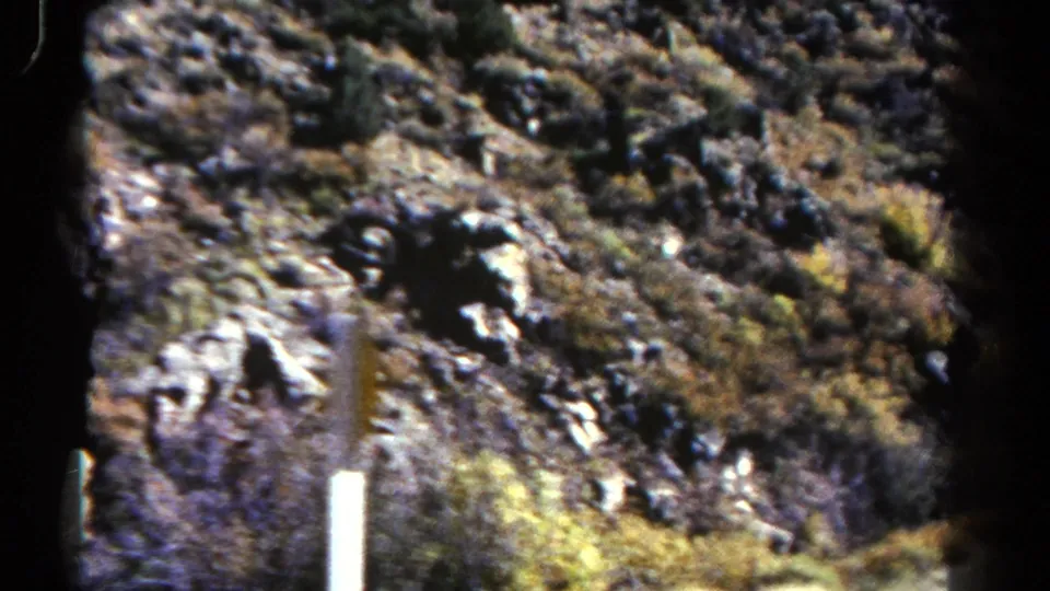 1967 Colorado travel frame with rugged hillside, scrub growth and vertical roadside posts viewed from inside a bus.