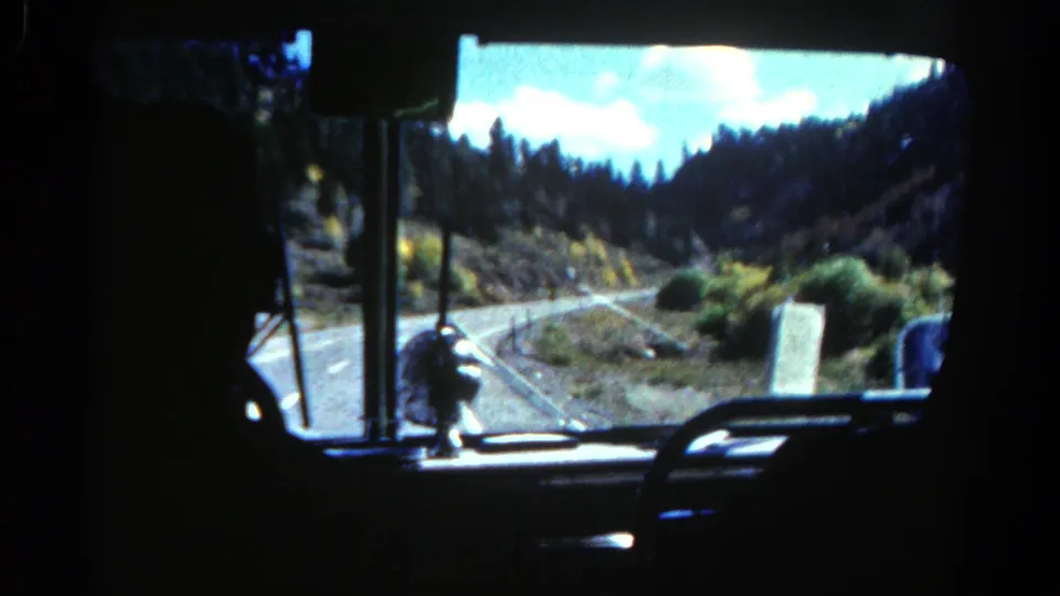 Interior bus view in 1967 Colorado showing a winding road, windshield frame and mountain slopes ahead.