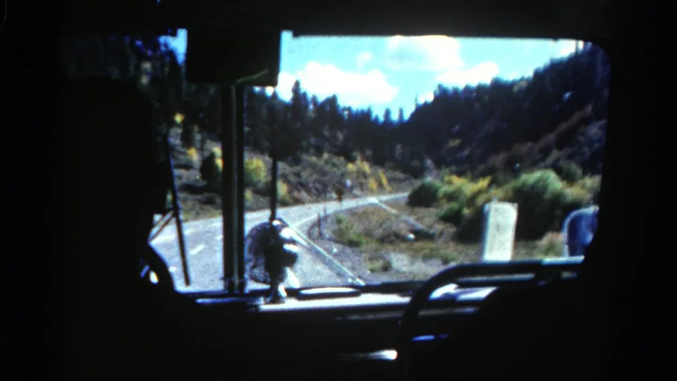 1967 Colorado 8mm view from inside a tour bus, looking through the windshield toward a curving road and pine-covered hills.
