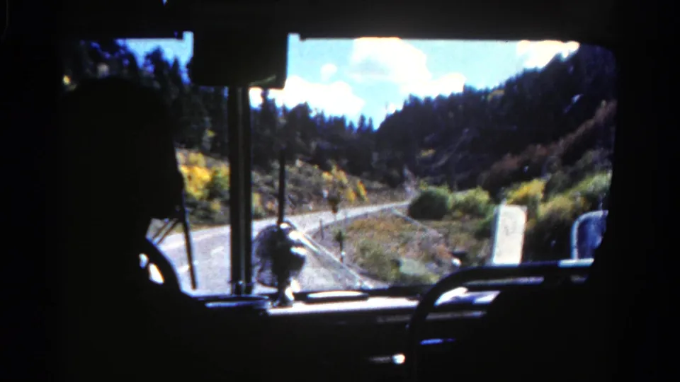 1967 Colorado home-movie frame with the tour bus driver in silhouette and a forest road visible through the windshield.