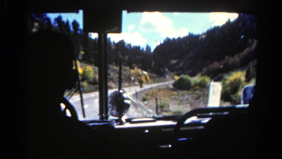 Colorado 1967 tour bus windshield view with dark interior framing a winding road, trees, and mountain hillside.