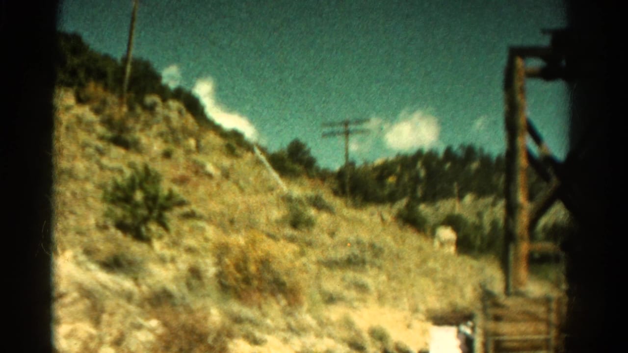 1967 Colorado roadside view with dry scrub-covered slope, utility poles, and a wooden structure near the edge of frame.