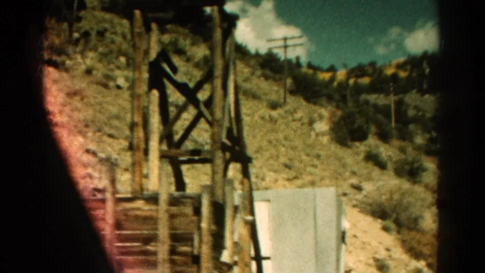 1967 Colorado 8mm frame passing a wooden roadside structure, utility poles, scrubby hillside, and pale service building.