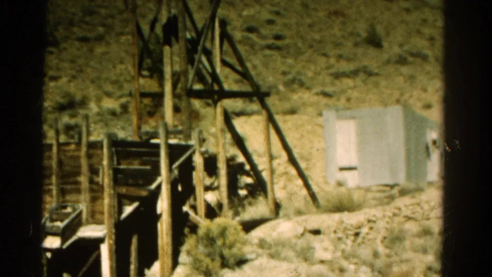 1967 Colorado roadside frame showing a weathered wooden structure, small light-colored building, rocky ground, and sparse brush.