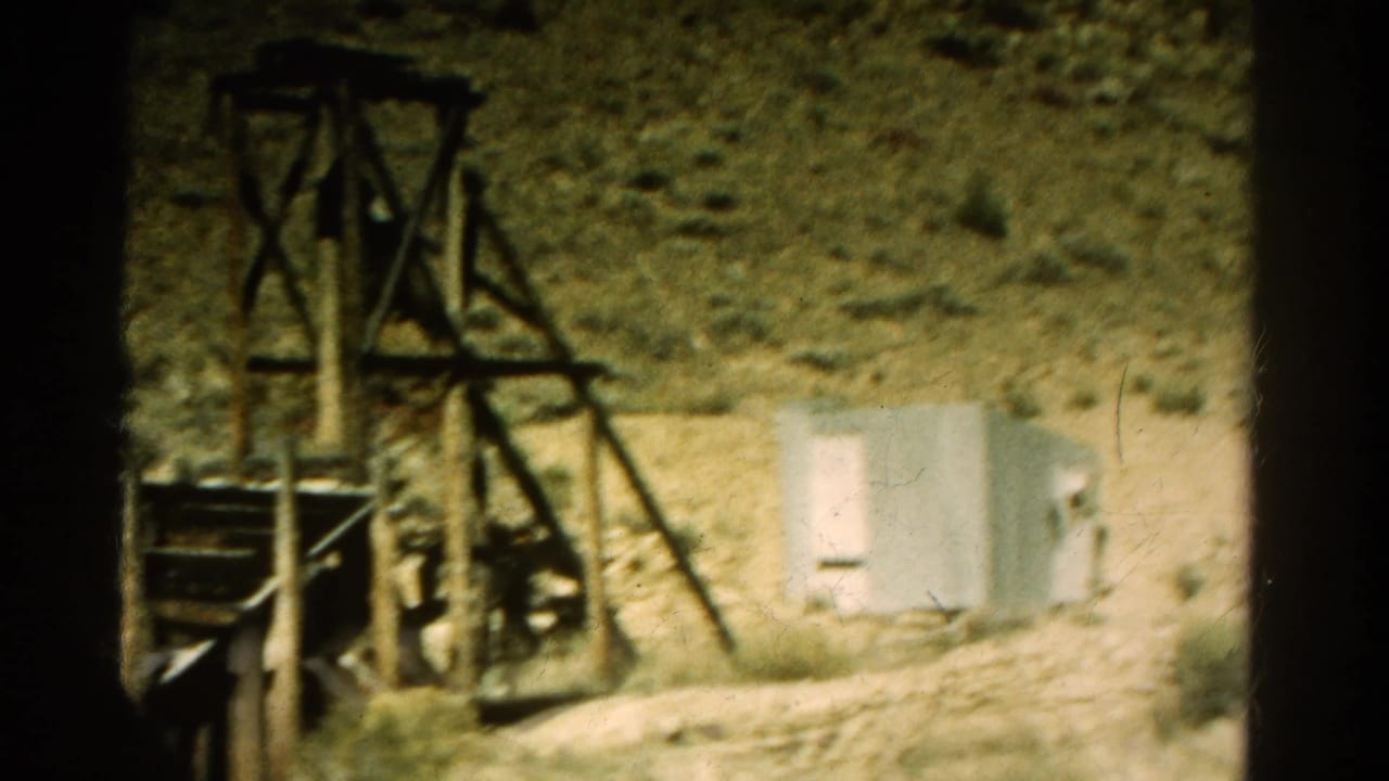 1967 Colorado home-movie frame showing a rough wooden structure beside a small shed in an arid hillside area.