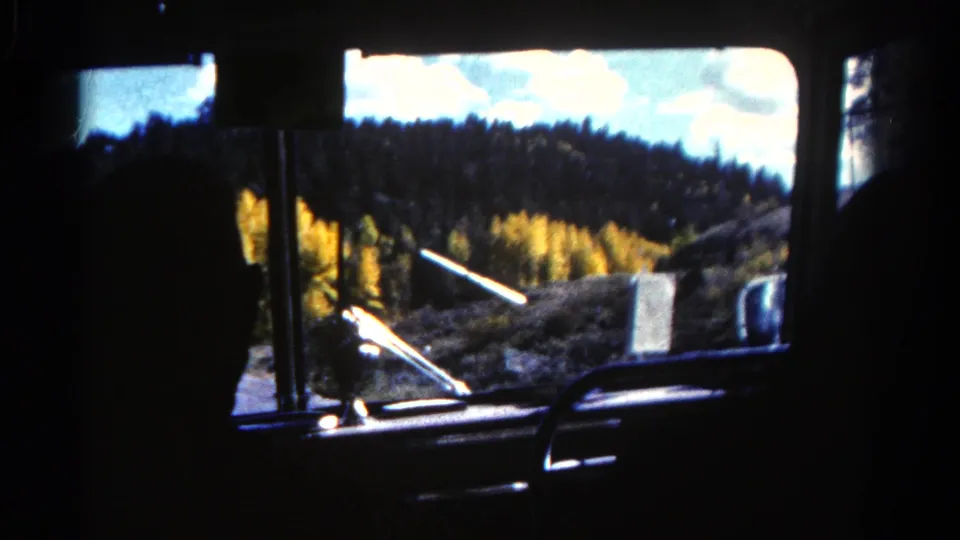Interior car-window view in 1967 Colorado, with silhouetted seats and wooded hills visible outside.