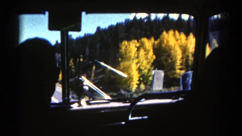 1967 home-movie frame from inside a moving vehicle, looking out at golden foliage and dark wooded hills in Colorado.