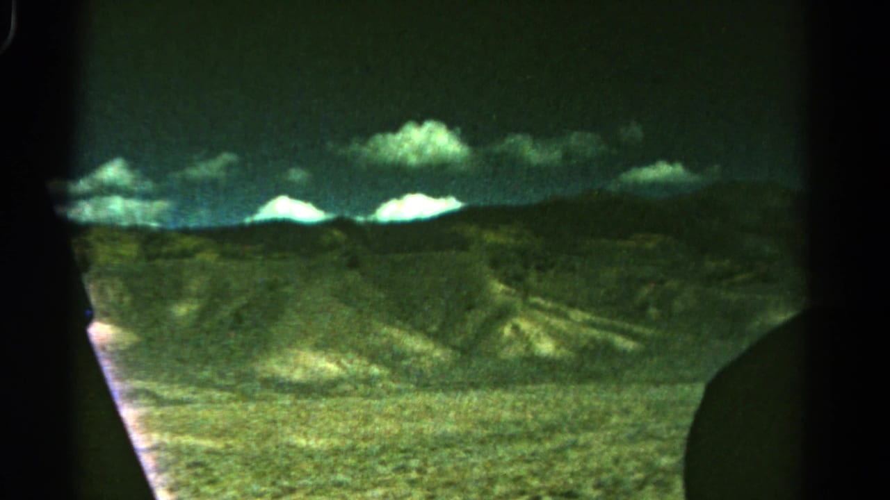 1967 Colorado home-movie frame with low mountain ridges, bright cloud forms, and open dry terrain viewed from a vehicle.