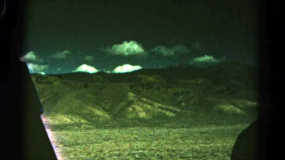 1967 Colorado 8mm frame with distant mountain silhouettes, bright clouds, and foreground scrubby ground filmed from a moving vehicle.