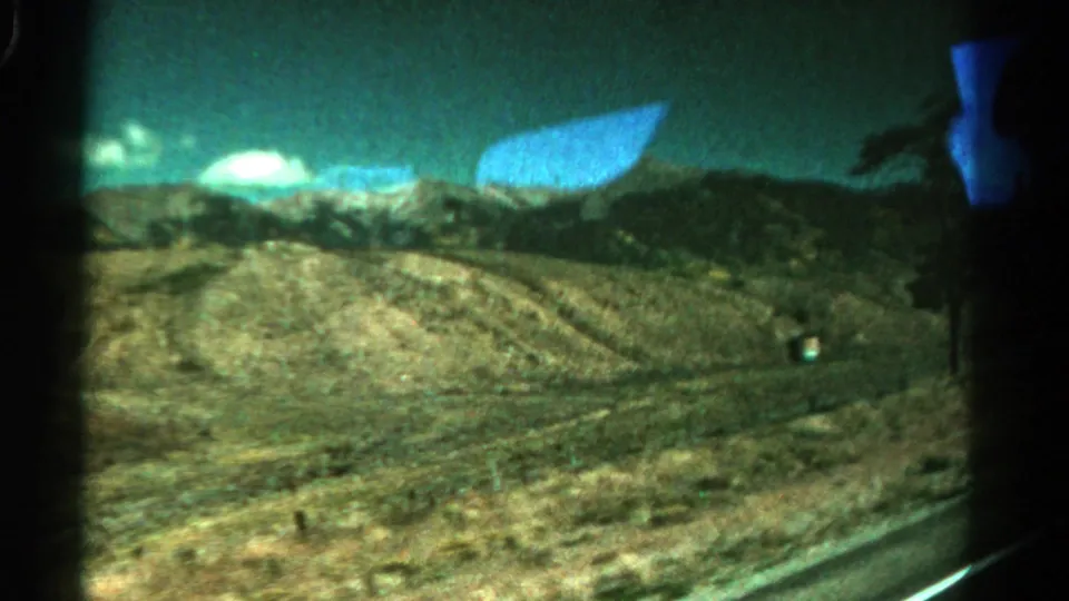 1967 Colorado road footage looking past dry hills and a roadside area, with a blue window reflection visible.