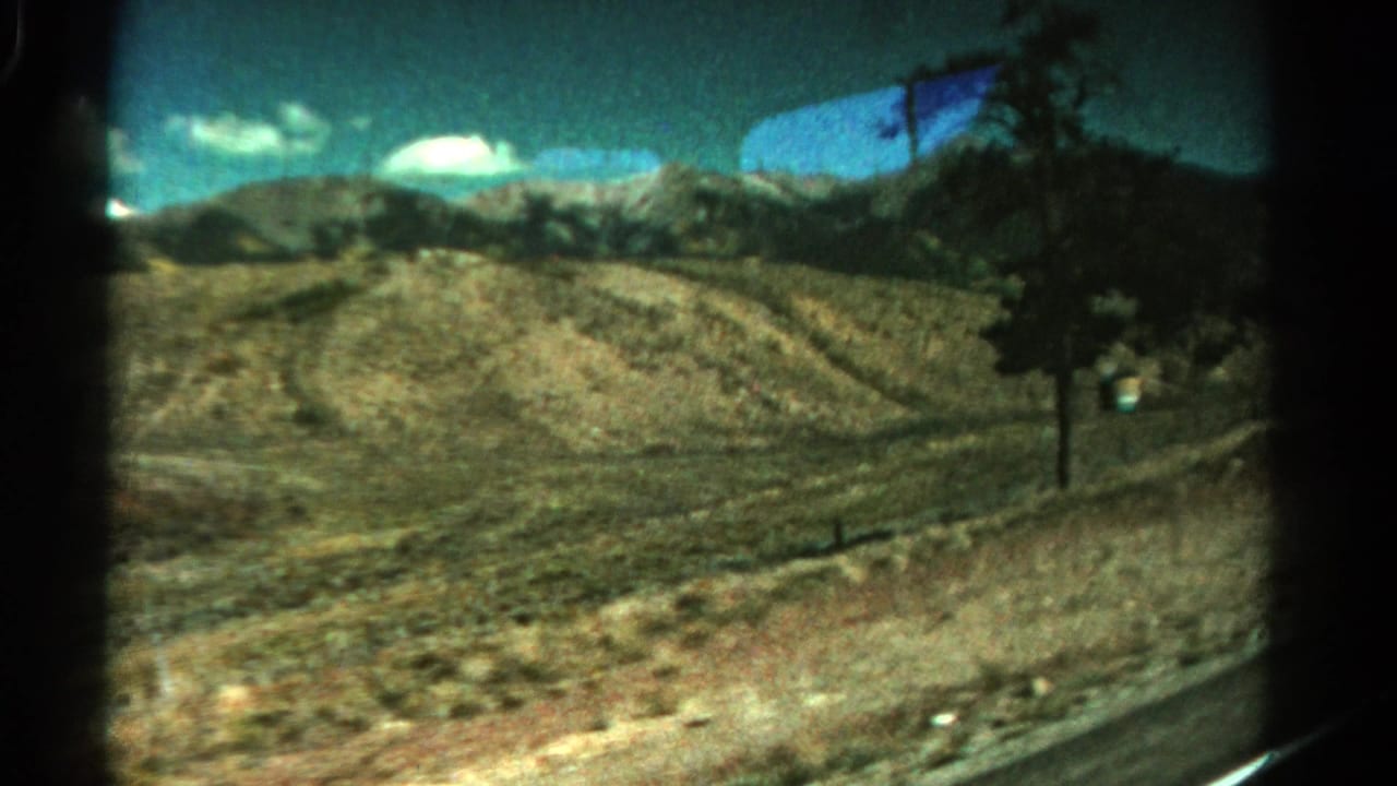 1967 Colorado 8mm frame showing dry rolling hills, a roadside tree, distant mountains, and blue sky from a moving vehicle.