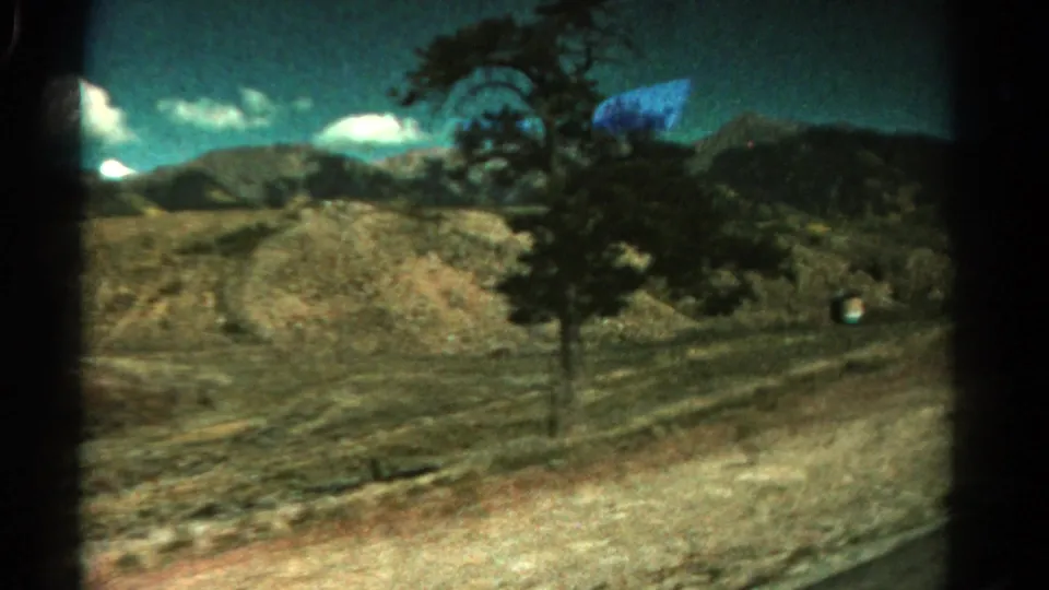 1967 Colorado home-movie frame with an open arid hillside, roadside tree, mountain ridge, and scattered clouds viewed from a vehicle.
