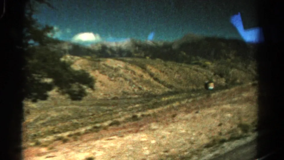 1967 Colorado 8mm frame showing a dry roadside slope, scattered trees, distant hills, and a blue reflection near the upper edge.